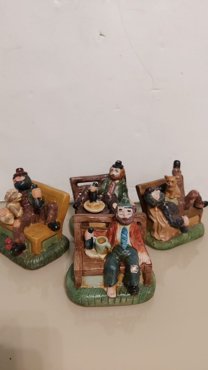 May include: Four ceramic figurines of men sitting on park benches. Each figurine is wearing different clothing and has a different expression. One figurine is holding a beer, another is holding a cup, and another is holding a cat. The figurines are painted in realistic colors and details.