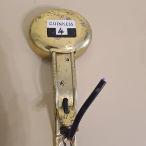 May include: A vintage brass Guinness tap handle with the number 4 on a black and white label. The handle shows signs of wear and tear, with a black cable attached. The tap is mounted on a beige wall.