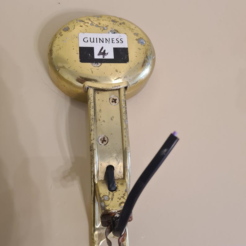 May include: A vintage brass Guinness tap handle with the number 4 on a black and white label. The handle shows signs of wear and tear, with a black cable attached. The tap is mounted on a beige wall.