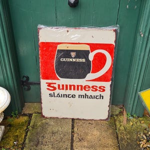 May include: A vintage metal sign advertising Guinness beer. The sign features a red background with a white outline of a pint glass filled with Guinness. The glass has the Guinness logo and the text "Guinness" in black. The sign also includes the text "Sláinte mhaith" in black.