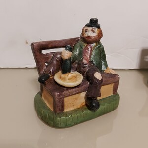 May include: A ceramic figurine of a man with a beard sitting on a bench with a beer and a plate of food. The figurine is painted in brown, green, and yellow.