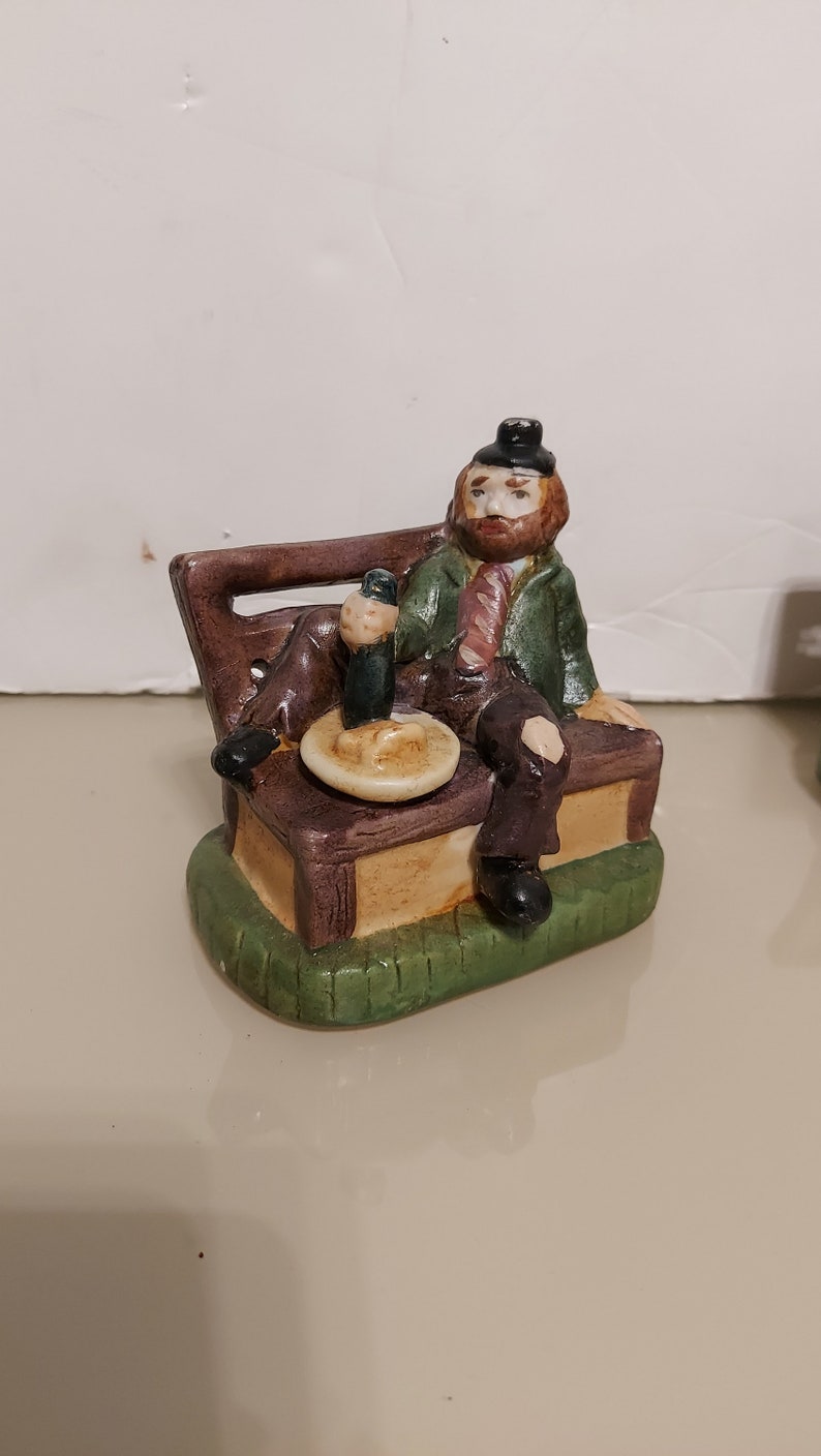 May include: A ceramic figurine of a man with a beard sitting on a bench with a beer and a plate of food. The figurine is painted in brown, green, and yellow.