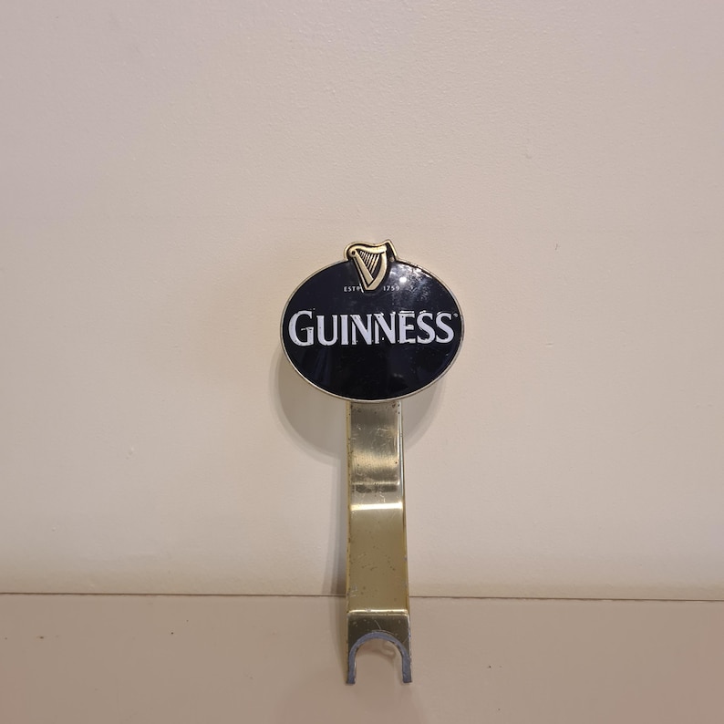 Puede incluir: Un grifo de cerveza Guinness con un letrero ovalado negro con el logotipo de Guinness en letras blancas. El grifo es de color dorado y tiene una base curvada. La parte superior del letrero tiene un s&iacute;mbolo de arpa dorada.