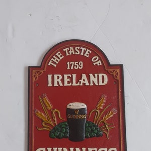 May include: A red and white cast iron sign with the text "THE TASTE OF IRELAND 1759 GUINNESS". The sign features a pint of Guinness, wheat stalks, and hops.