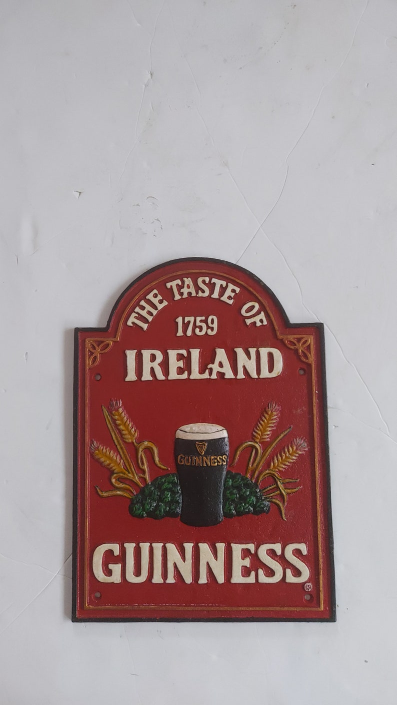 May include: A red and white cast iron sign with the text "THE TASTE OF IRELAND 1759 GUINNESS". The sign features a pint of Guinness, wheat stalks, and hops.