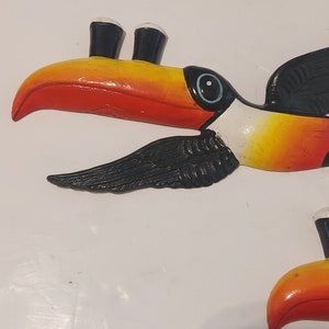 Op de afbeelding: Een kleurrijke toekan sculptuur met een grote oranje en gele snavel, zwarte vleugels en een wit oog. De vogel heeft een zwart-witte kop en is geschilderd op een wit oppervlak. De toekan is een tropische vogel.