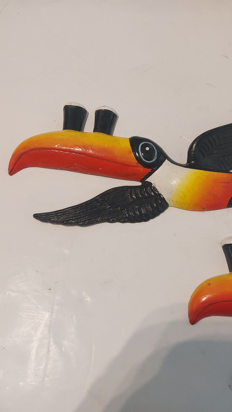 Op de afbeelding: Een kleurrijke toekan sculptuur met een grote oranje en gele snavel, zwarte vleugels en een wit oog. De vogel heeft een zwart-witte kop en is geschilderd op een wit oppervlak. De toekan is een tropische vogel.