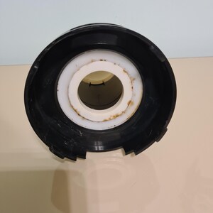 May include: Black plastic lid with a white plastic ring and a central hole. The lid has a textured surface and appears to be for a container.