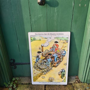 K&ouml;nnte beinhalten: Ein Vintage-Metall-Schild mit einer Cartoon-Illustration einer dampfbetriebenen Maschine, die Gerste auf einem Feld erntet. Das Schild tr&auml;gt die Aufschrift "The Gentle Art of Making Guinness. Early Morning On The Guinness Farm."