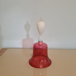 Può includere: Una campana di vetro rosa con una parte superiore in vetro bianco. La campana è appoggiata su una superficie marrone chiaro.