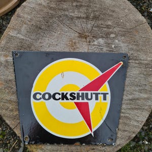 May include: A black and yellow enamel sign with a red arrow pointing to the right. The sign says "COCKSSHUTT" in white letters.