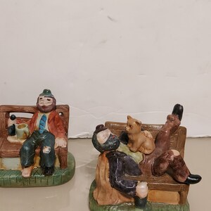 May include: Two ceramic figurines of men sitting on benches. The man on the left is wearing a green hat, a red jacket, and a blue tie. He is holding a beer mug. The man on the right is wearing a black jacket and has a cat sitting on his lap. Both figurines are painted in realistic detail.