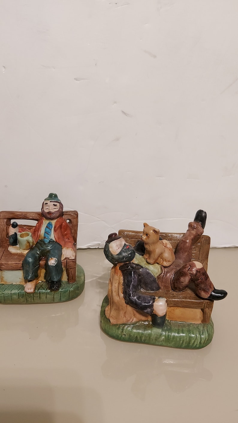 May include: Two ceramic figurines of men sitting on benches. The man on the left is wearing a green hat, a red jacket, and a blue tie. He is holding a beer mug. The man on the right is wearing a black jacket and has a cat sitting on his lap. Both figurines are painted in realistic detail.