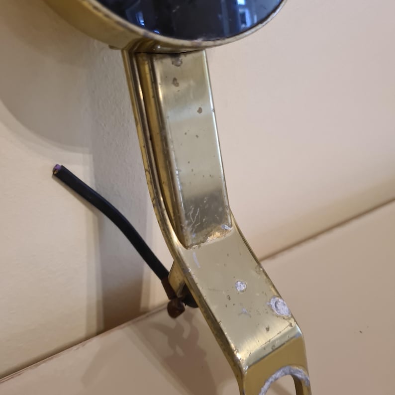 May include: Close-up of a vintage brass-toned vanity mirror with a circular reflective surface. The mirror's frame shows signs of wear, with visible scratches and discoloration. A black wire extends from the base, suggesting an electrical component.