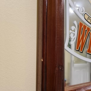 Puede incluir: Una secci&oacute;n de un espejo con marco de madera y acabado marr&oacute;n oscuro. El espejo tiene un dise&ntilde;o decorativo con la palabra "W" en letras naranjas y blancas. El espejo est&aacute; montado en una pared de color beige claro.