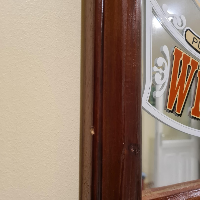 Puede incluir: Una secci&oacute;n de un espejo con marco de madera y acabado marr&oacute;n oscuro. El espejo tiene un dise&ntilde;o decorativo con la palabra "W" en letras naranjas y blancas. El espejo est&aacute; montado en una pared de color beige claro.