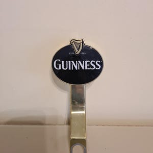 Puede incluir: Un grifo de cerveza Guinness dorado con un emblema ovalado negro. El emblema muestra el logotipo de Guinness en blanco, junto con un s&iacute;mbolo de arpa. Tambi&eacute;n es visible el texto 'ESTD 1759'.