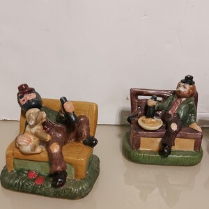 May include: Two ceramic figurines of men sitting on benches. The man on the left is holding a bottle and has a dog on his lap. The man on the right is holding a glass and a plate. Both figurines are wearing hats and have facial hair.