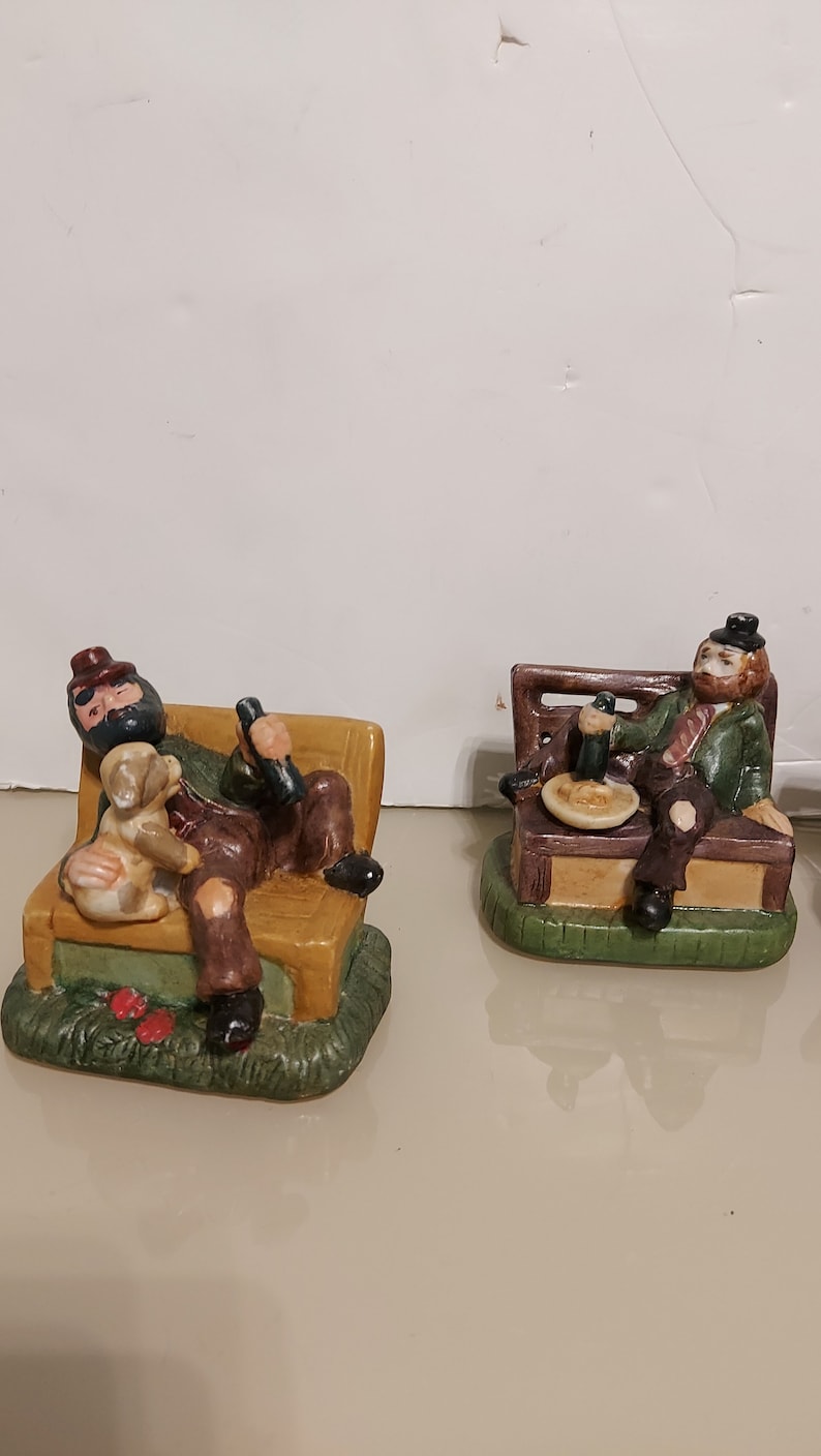 May include: Two ceramic figurines of men sitting on benches. The man on the left is holding a bottle and has a dog on his lap. The man on the right is holding a glass and a plate. Both figurines are wearing hats and have facial hair.