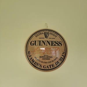 May include: A round Guinness sign with a brown background and black and red text. The sign features the Guinness logo, harp symbol, and the text "St. James's Gate Dublin". The sign is hanging on a light yellow wall.