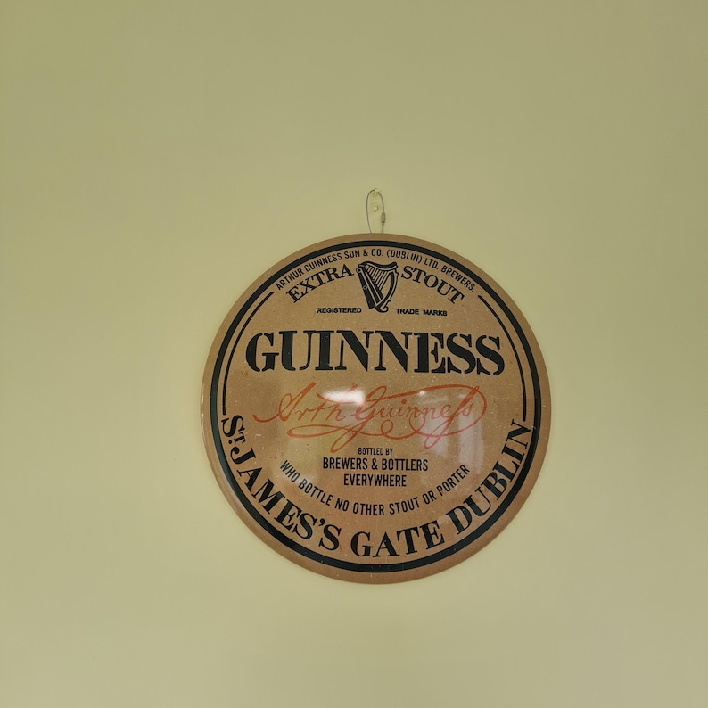 May include: A round Guinness sign with a brown background and black and red text. The sign features the Guinness logo, harp symbol, and the text "St. James's Gate Dublin". The sign is hanging on a light yellow wall.