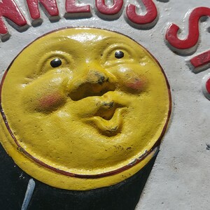 May include: A vintage cast iron sign with a yellow smiling moon face and the word "NESS" in red letters.