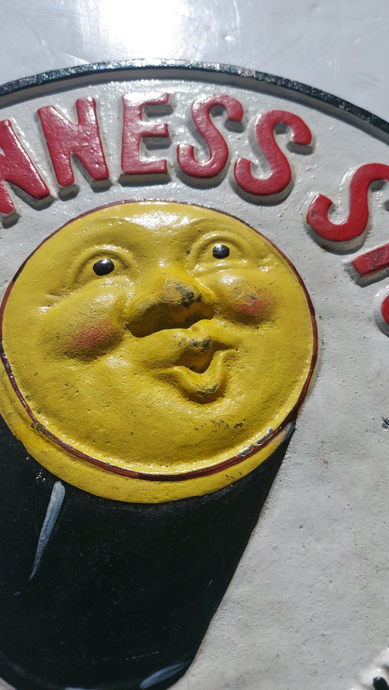 May include: A vintage cast iron sign with a yellow smiling moon face and the word "NESS" in red letters.