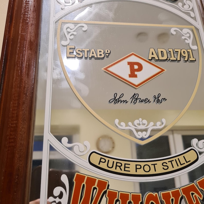 Puede incluir: Espejo vintage con marco de madera marr&oacute;n. El espejo presenta el texto "ESTAB? AD.1791" y "PURE POT STILL". Tambi&eacute;n es visible un logotipo en forma de diamante con la letra "P". El espejo tiene un borde decorativo.