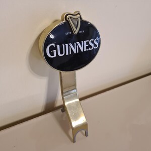 Puede incluir: Un tirador de grifo Guinness de lat&oacute;n con un emblema ovalado en blanco y negro. El emblema presenta el logotipo de Guinness y el texto "ESTD 1759". El tirador est&aacute; dise&ntilde;ado para dispensar cerveza negra Guinness.