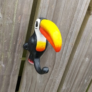 May include: A black toucan-shaped hook with a yellow beak, orange and white accents, mounted on a wooden fence.