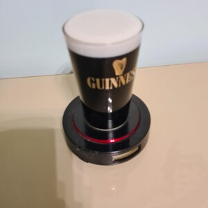 May include: A black and gold Guinness pint glass with a white head of foam, sitting on a black rotating base with a red ring.