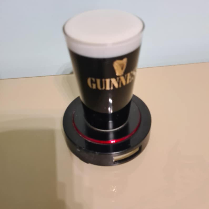 May include: A black and gold Guinness pint glass with a white head of foam, sitting on a black rotating base with a red ring.