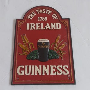 May include: A red and gold cast iron sign with the text "THE TASTE OF IRELAND 1759" and "GUINNESS". The sign features a pint of Guinness, wheat stalks, and hops.