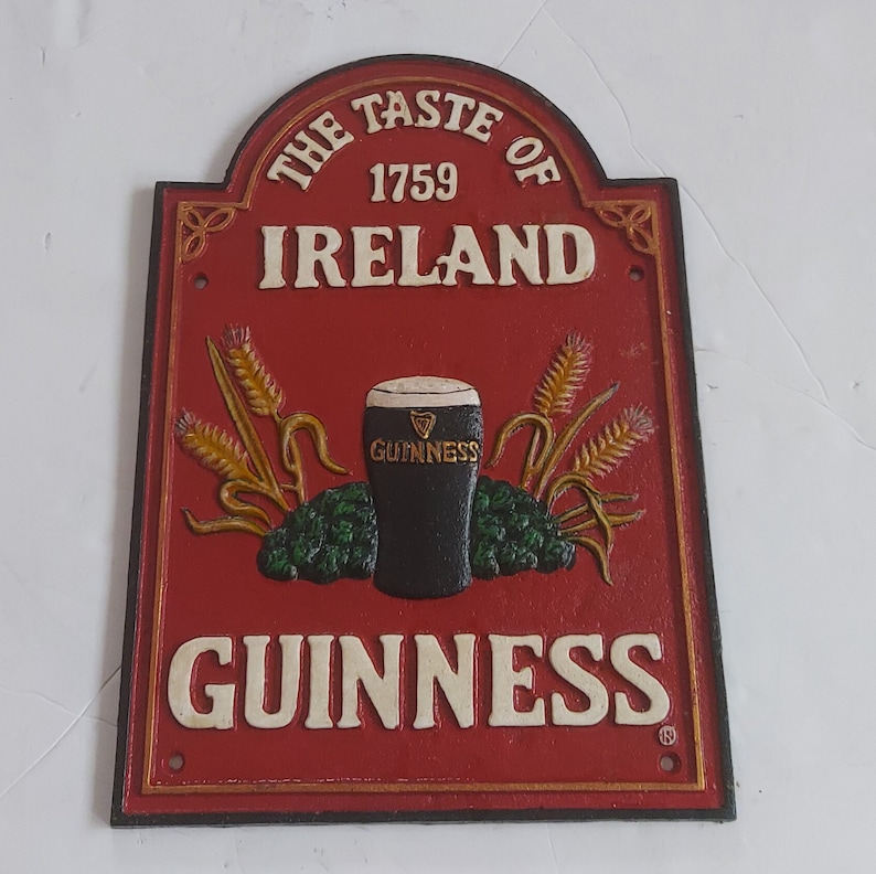 May include: A red and gold cast iron sign with the text "THE TASTE OF IRELAND 1759" and "GUINNESS". The sign features a pint of Guinness, wheat stalks, and hops.