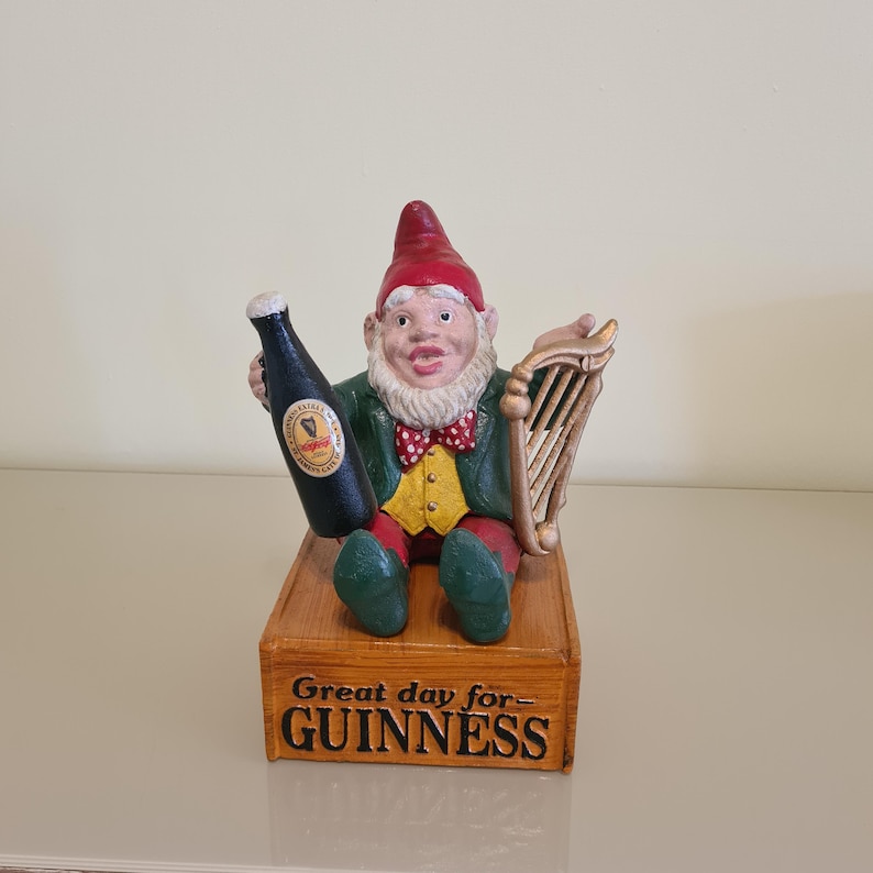 May include: A whimsical garden gnome figurine holding a Guinness bottle and a harp. The gnome wears a red hat, green jacket, and a bow tie. The base reads "Great day for Guinness."