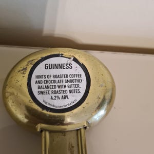 Puede incluir: Un abrebotellas dorado con la palabra "GUINNESS" y texto que describe el perfil de sabor de la cerveza: caf&eacute; tostado, chocolate, amargo, dulce y notas tostadas. El abridor tambi&eacute;n indica 4,2% ABV.