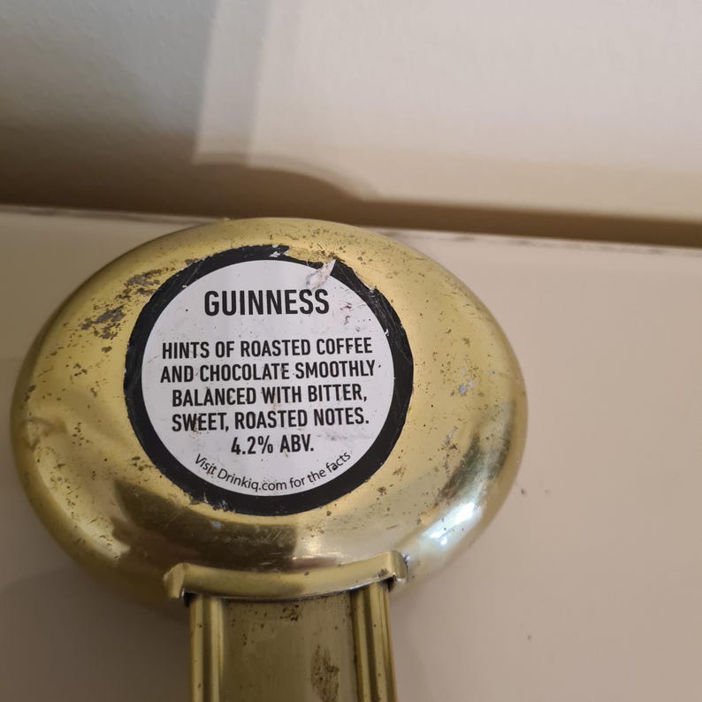 Puede incluir: Un abrebotellas dorado con la palabra "GUINNESS" y texto que describe el perfil de sabor de la cerveza: caf&eacute; tostado, chocolate, amargo, dulce y notas tostadas. El abridor tambi&eacute;n indica 4,2% ABV.