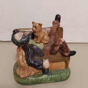 May include: A ceramic figurine of a man sleeping on a bench with a small dog sitting on his lap. The man is wearing a brown jacket and a black hat. The bench is brown and has a green base.