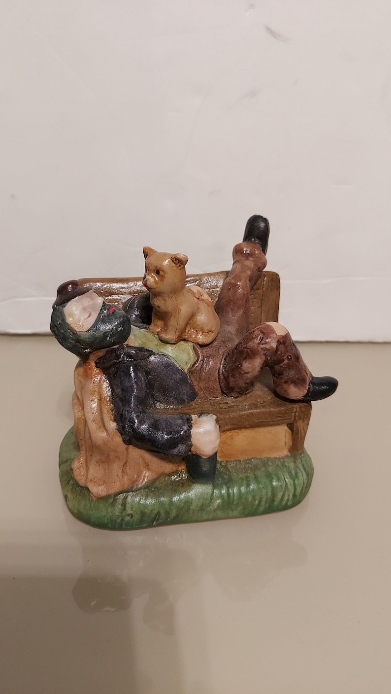 May include: A ceramic figurine of a man sleeping on a bench with a small dog sitting on his lap. The man is wearing a brown jacket and a black hat. The bench is brown and has a green base.