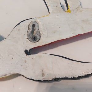 Op de afbeelding: Een wit, handgeschilderd pelikaan wanddecoratie met zwarte, rode en gele accenten. De houten vogel heeft een metalen hanger aan de achterkant. De bek van de pelikaan staat open en de vogel is geschilderd met een distressed afwerking.