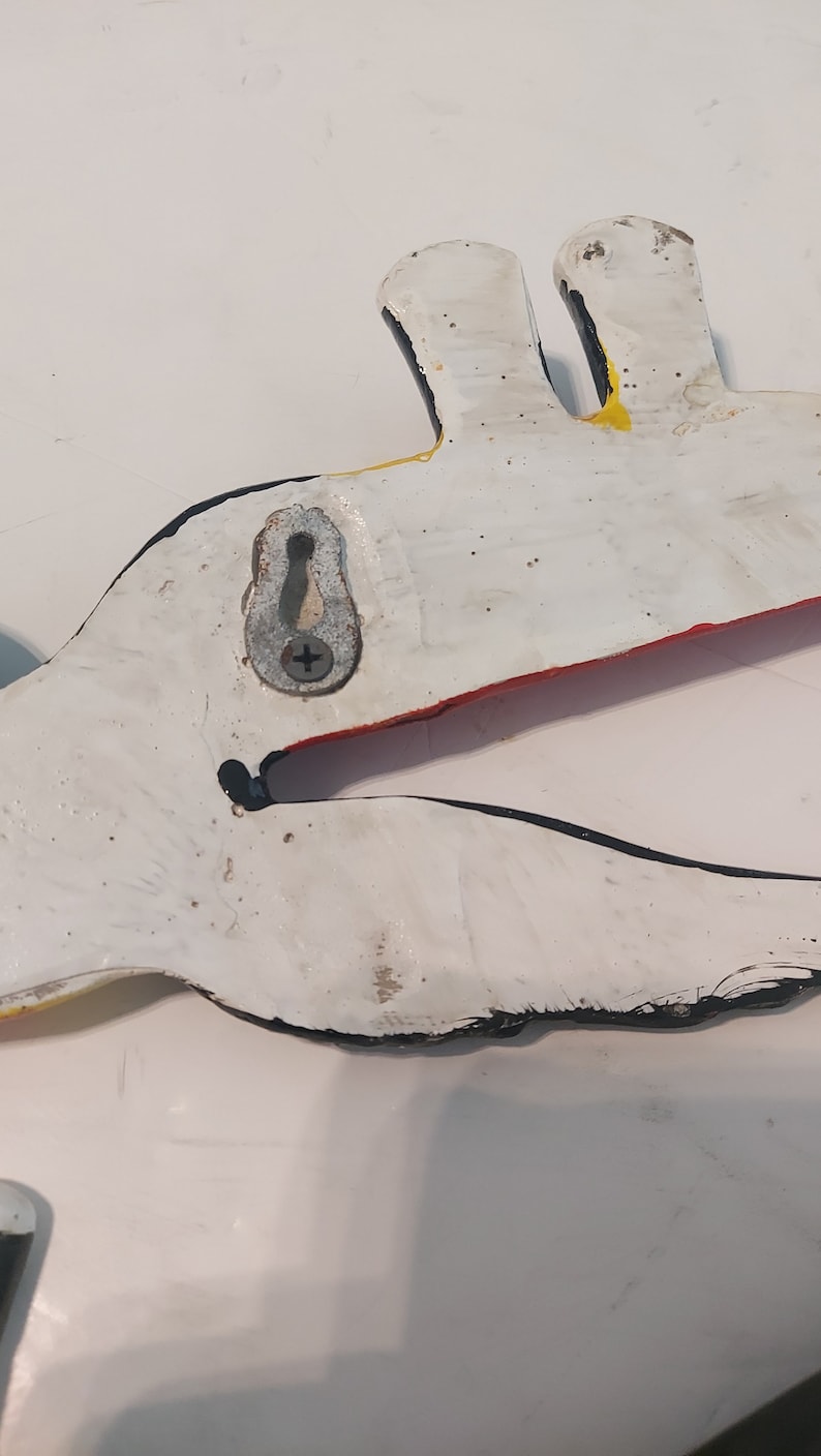 Op de afbeelding: Een wit, handgeschilderd pelikaan wanddecoratie met zwarte, rode en gele accenten. De houten vogel heeft een metalen hanger aan de achterkant. De bek van de pelikaan staat open en de vogel is geschilderd met een distressed afwerking.