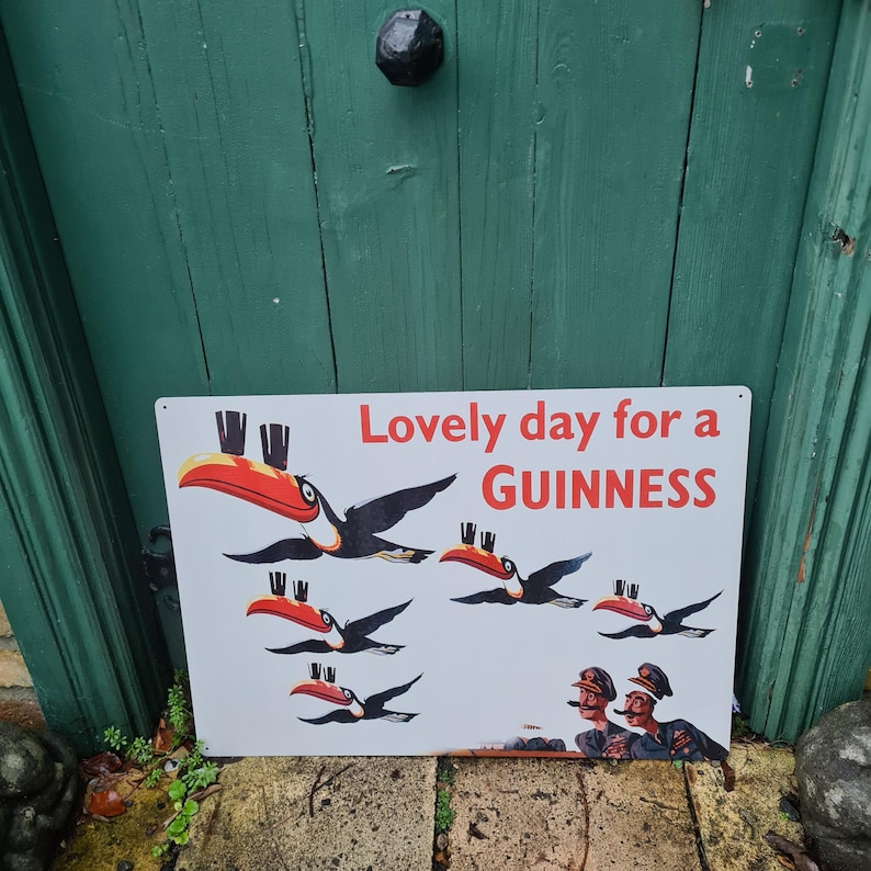 May include: A rectangular white metal sign with the text "Lovely day for a GUINNESS" in red. The sign features illustrations of toucans with red and yellow beaks and black wings. Two men in military uniforms are at the bottom.