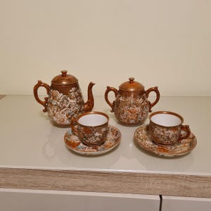Puede incluir: Un juego de té vintage con un diseño floral marrón. El juego incluye una tetera, un azucarero y dos tazas de té con platillos. La tetera mide aproximadamente 20 cm de altura.