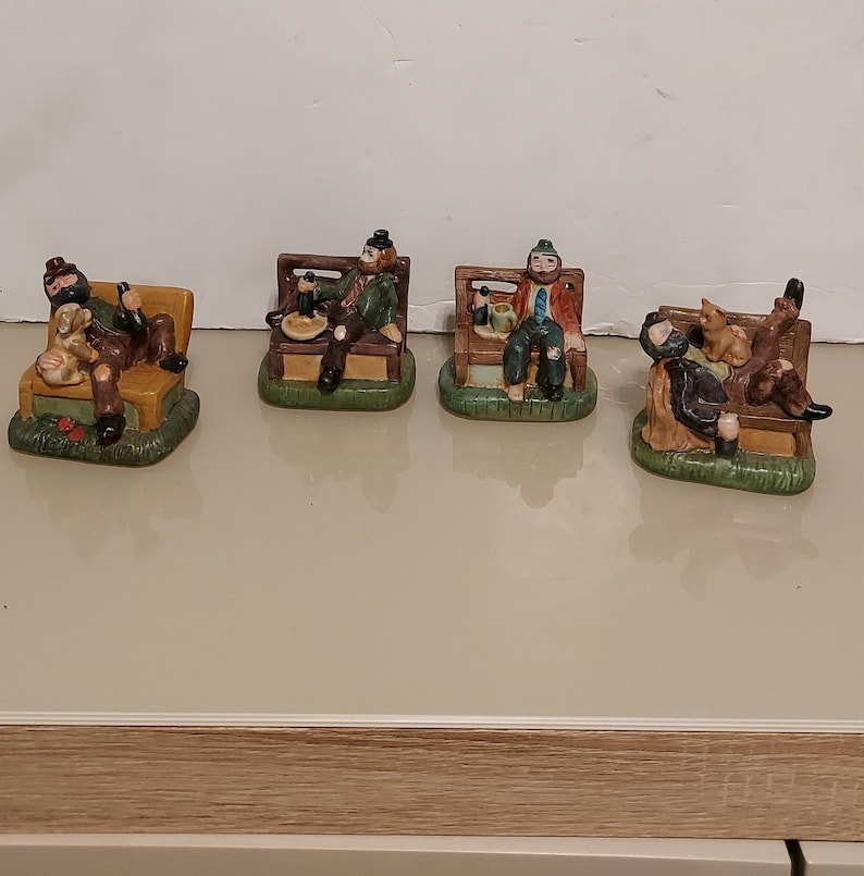 May include: Four ceramic figurines of men sitting on park benches. Each figurine is wearing a different outfit and has a different expression on their face. The figurines are painted in realistic detail and are a charming addition to any home decor.