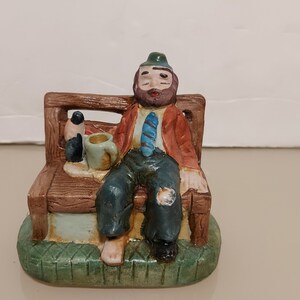 May include: A ceramic figurine of a man sitting on a bench with a bottle and a cup in his hands. He is wearing a red jacket, a blue tie, and a green hat. The figurine is painted in brown, green, blue, and red colors.