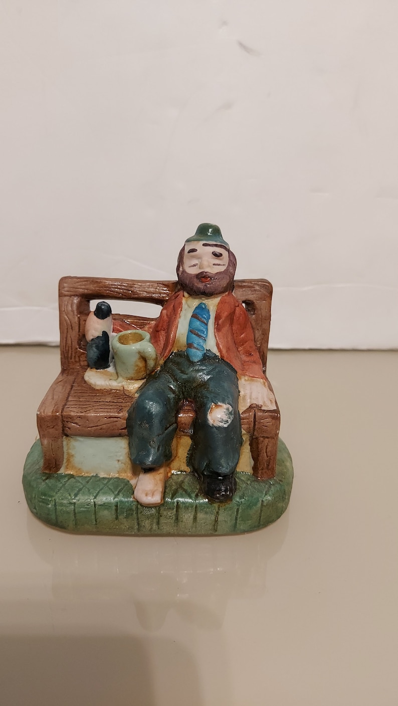 May include: A ceramic figurine of a man sitting on a bench with a bottle and a cup in his hands. He is wearing a red jacket, a blue tie, and a green hat. The figurine is painted in brown, green, blue, and red colors.