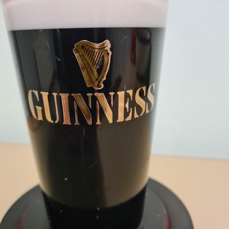May include: A black glass pint glass with a white head of foam. The glass has a gold Guinness logo with a harp design.