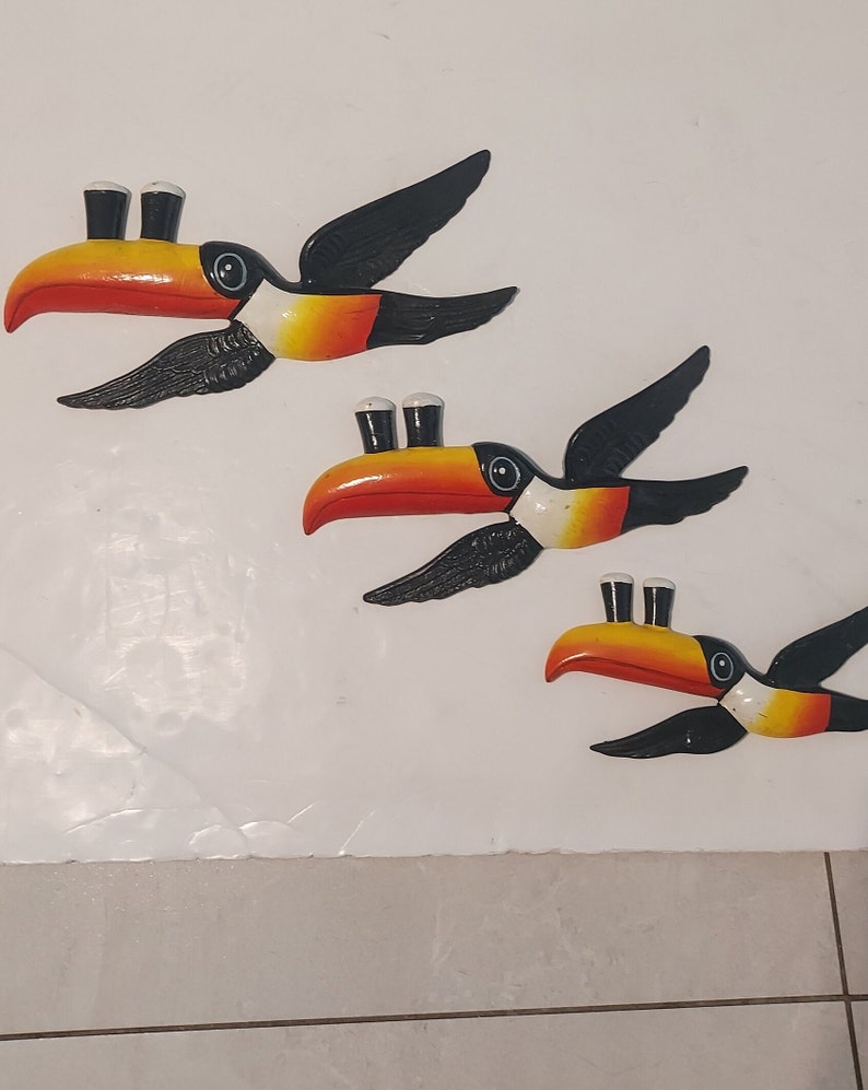 Op de afbeelding: Drie wanddecoraties van toekans. Elke toekan heeft een zwarte vleugel, een rode en gele snavel en een zwart-wit oog. De toekans hebben twee kleine zwarte cilinders bovenop hun hoofd.