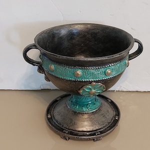 May include: A black and teal ceramic goblet with decorative details and a round base. The goblet has two handles and a wide, round bowl.