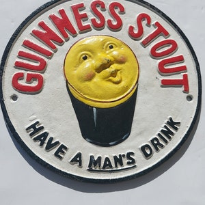 May include: A white, round, cast iron sign with a black border. The sign has red lettering that says "Guinness Stout" and "Have a Man's Drink". In the center of the sign is a yellow cartoon face with a black pint glass.
